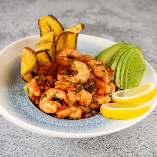 Shrimp ceviche with plantain chips, sliced avocado, and lemon wedges in a white and blue bowl.