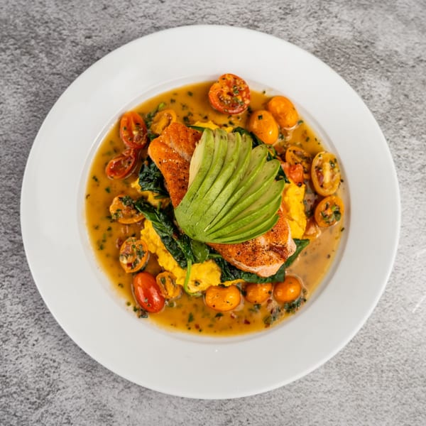 Seared salmon with sliced avocado, spinach, scrambled eggs, and cherry tomatoes in a savory sauce.